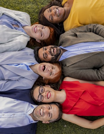 Group of multiracial business professionals lying on grass, smiling, and bonding during an outdoor team-building activity.